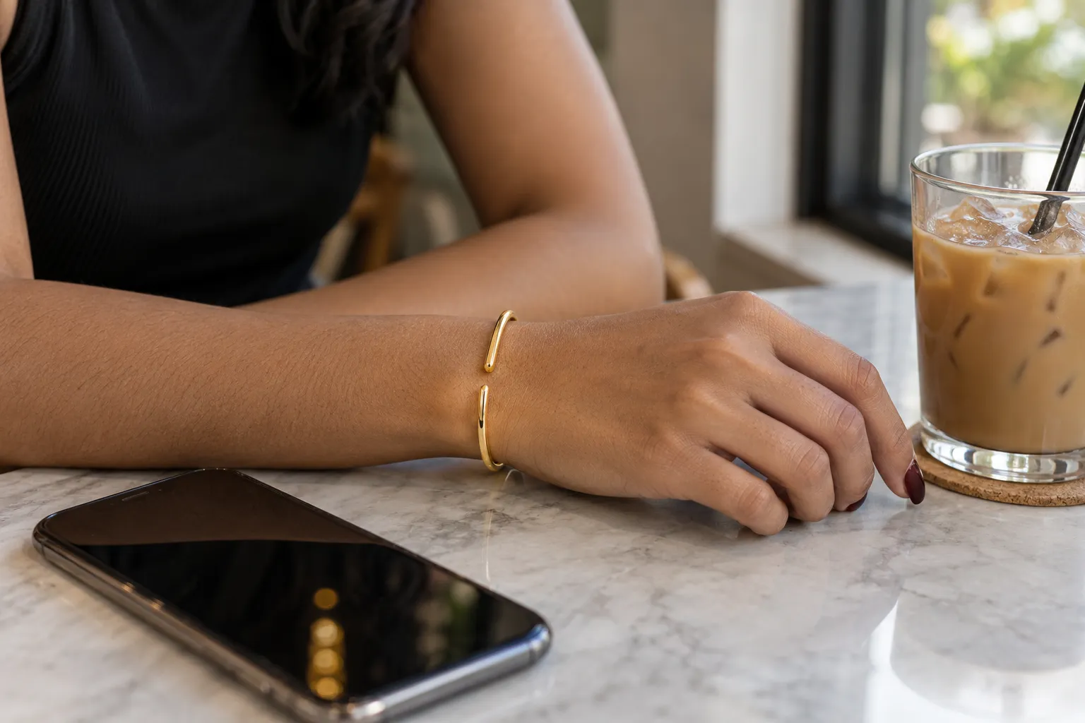 A slim gold cuff on a woman’s wrist at a café table, beside her phone