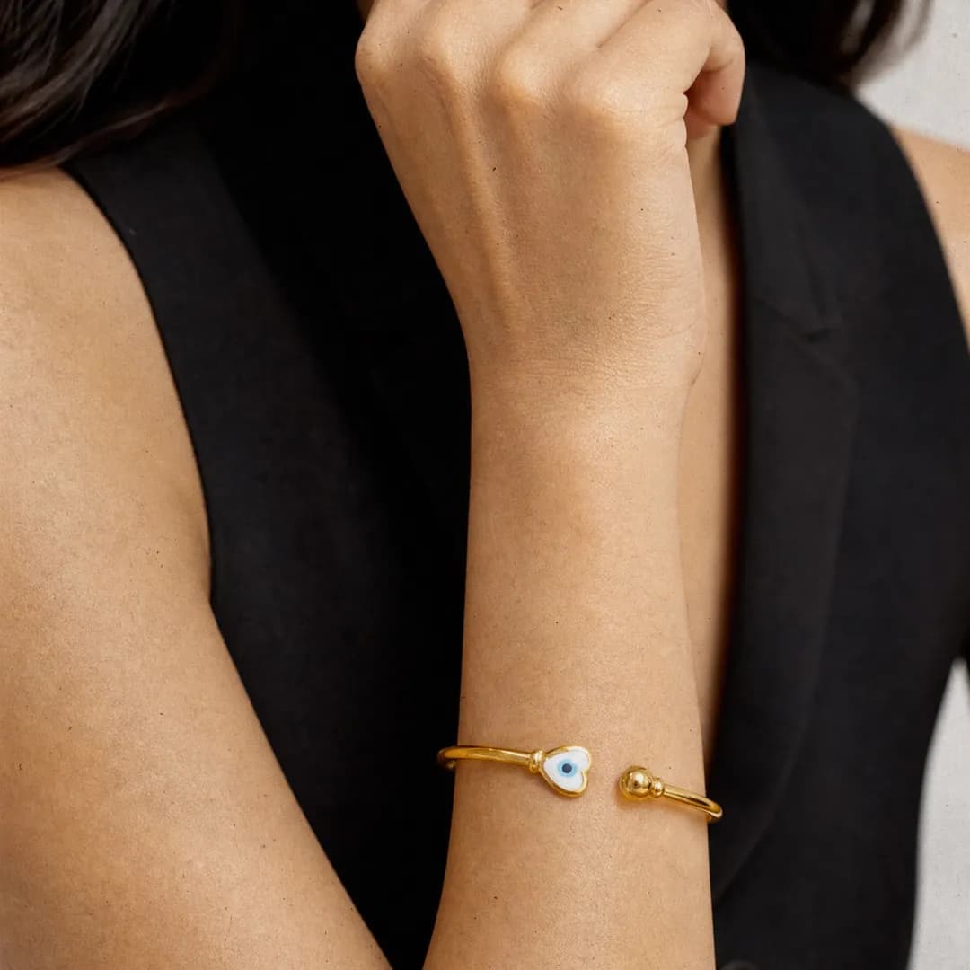 A slim gold open cuff with a heart-shaped evil eye charm, worn on the wrist