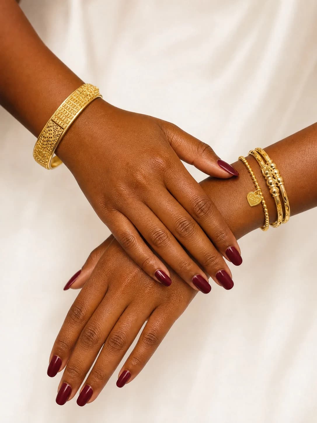 Two crossed hands wearing gold bangles, a wide filigree bangle on one wrist and slim bangles with a heart charm on the other