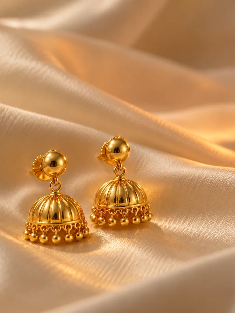 A pair of gold jhumka earrings on ivory silk beside a lit clay diya
